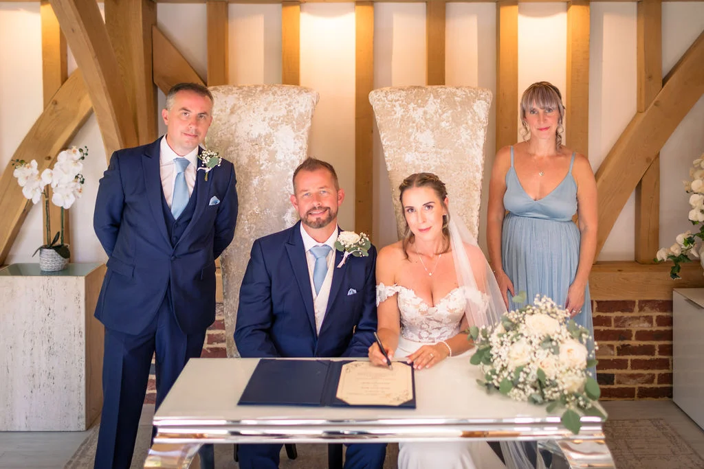 A bride signs the marriage register at a rustic barn venue alongside her groom, best man, and maid of honour.