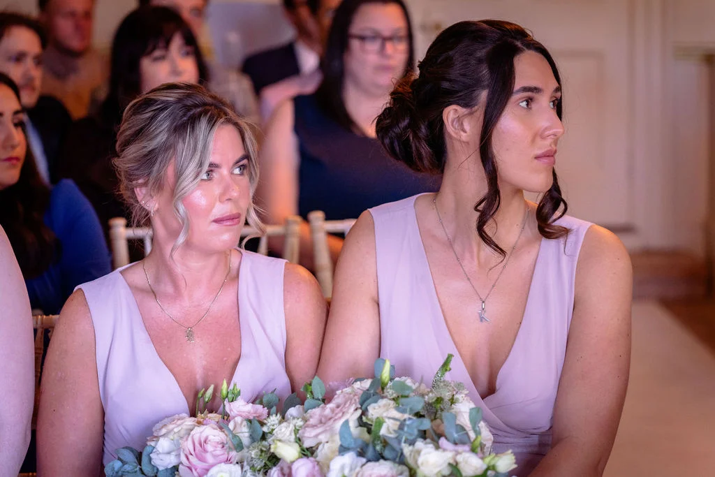 Two bridesmaids wearing matching pink V-neck dresses and delicate silver necklaces sit attentively during the wedding ceremony holding lush floral bouquets.