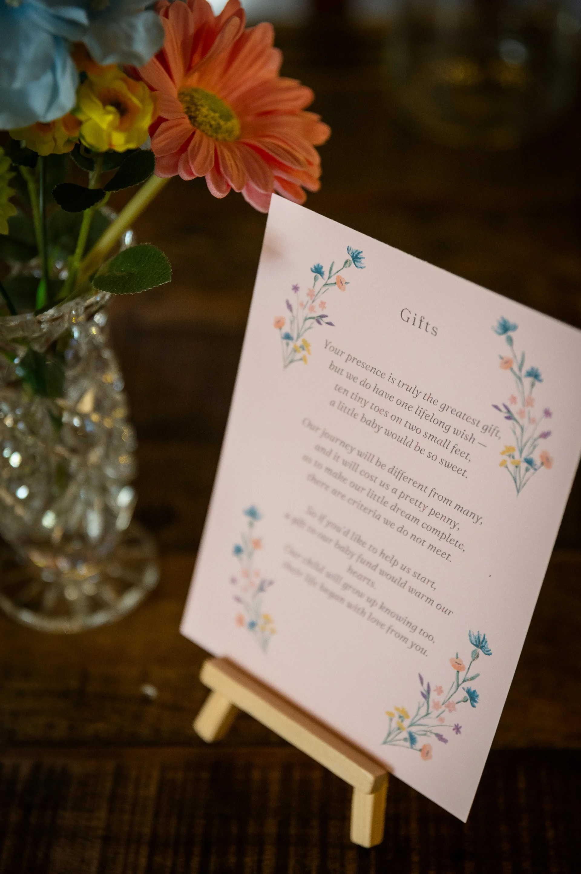 A close-up shot shows a custom printed gift sign displayed on a small wooden easel alongside a vintage glass vase filled with colourful wildflowers.