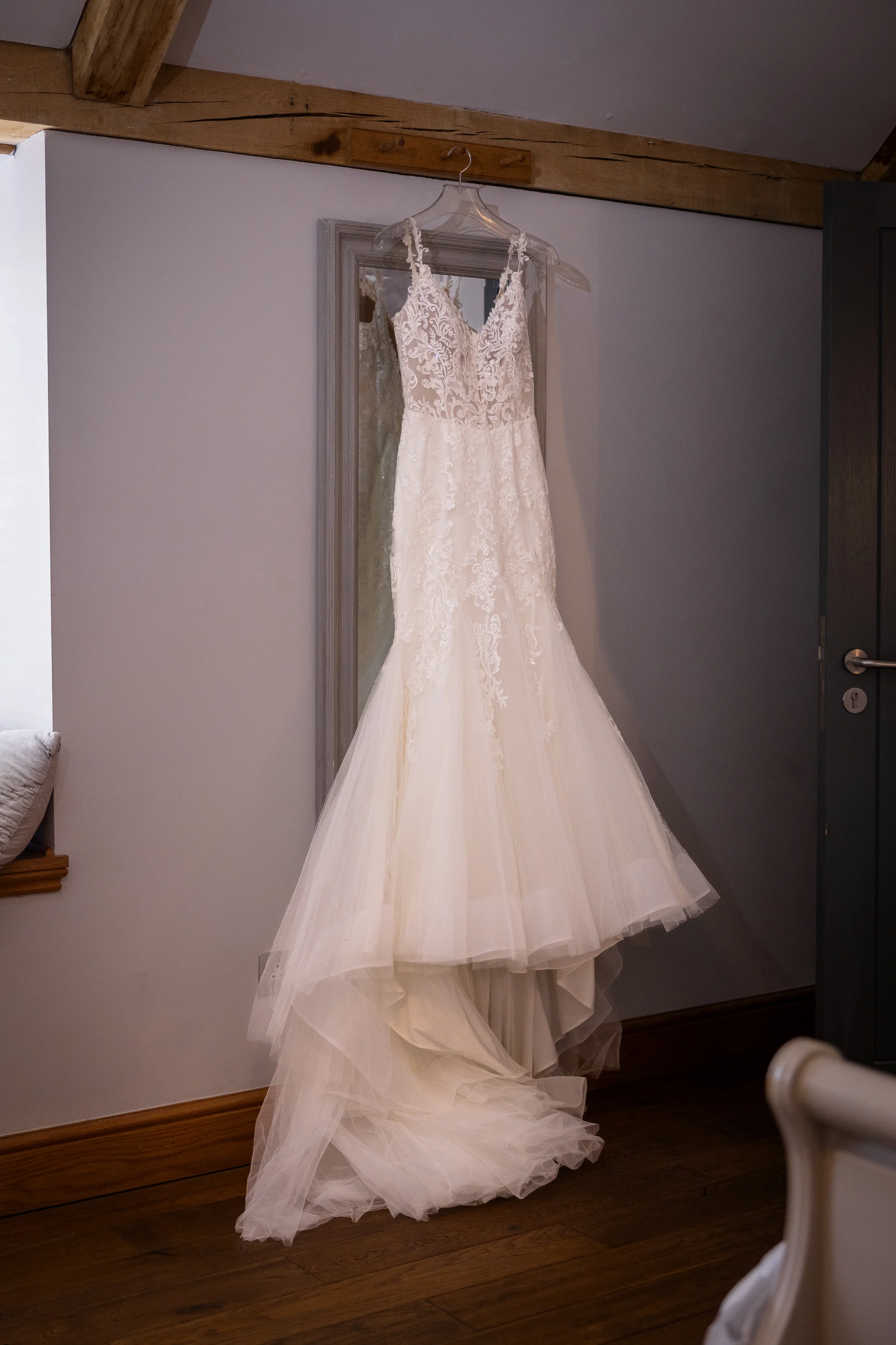 A delicate lace fit-and-flare wedding dress hangs neatly from a mirror in a light-filled room with wooden ceiling beams.