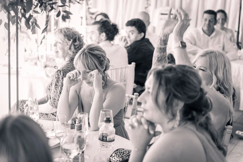 Wedding guests seated at a reception table laugh and wipe away tears of joy while listening to the speeches.
