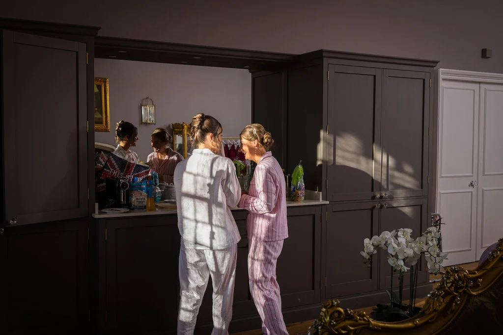 Two women wearing matching pyjamas stand chatting in a kitchen while getting ready for a wedding, illuminated by bright sunlight streaming through a window.