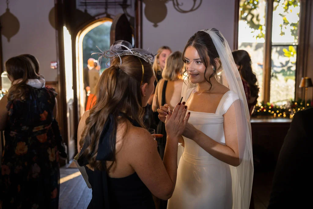 A radiant bride in a sleek white satin gown with a square neckline and veil smiles warmly while chatting with a guest during a drinks reception at a stately home.