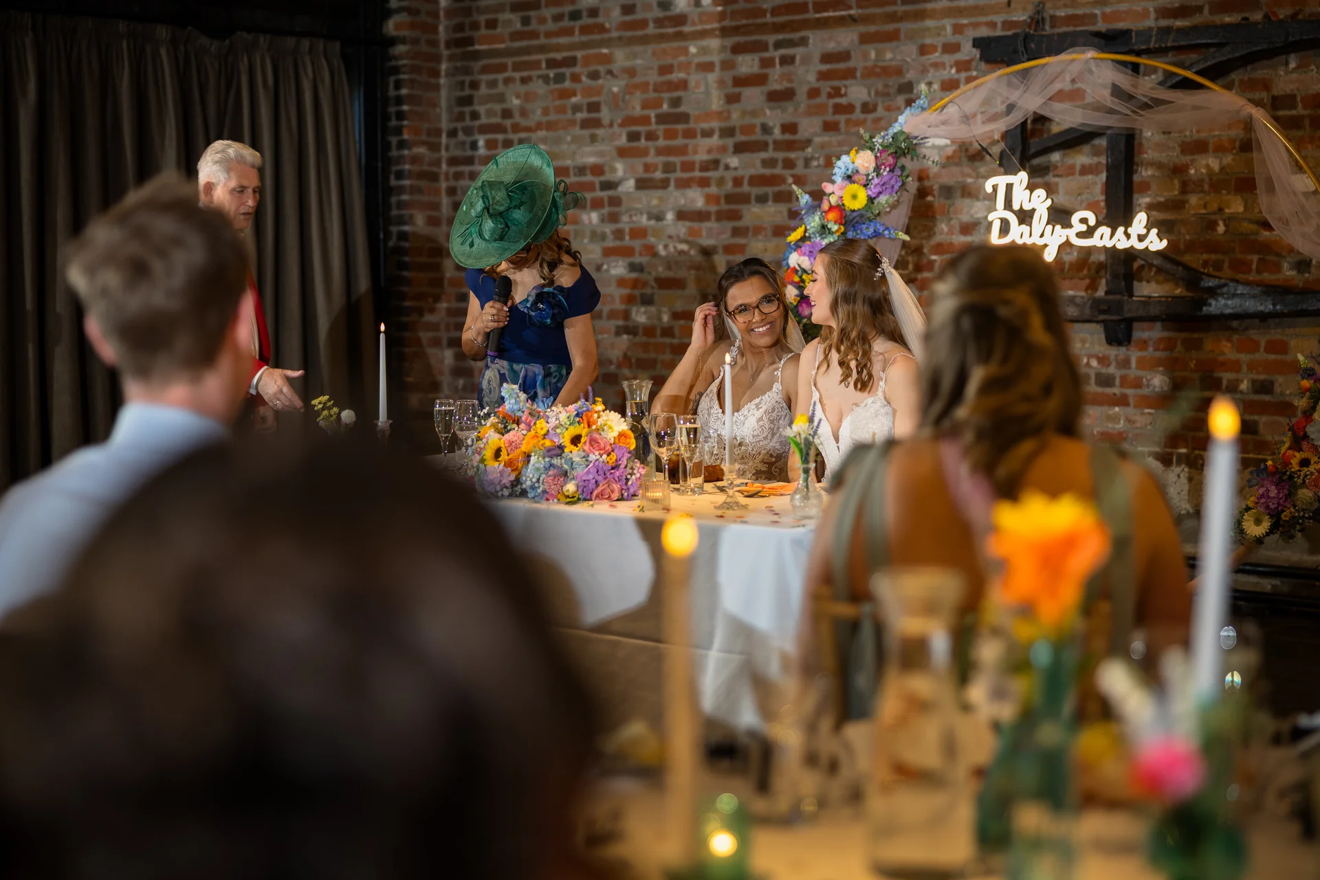 Two smiling brides sit at the top table during wedding speeches, with a guest in a dramatic green hat and fascinator speaking into a microphone nearby.
