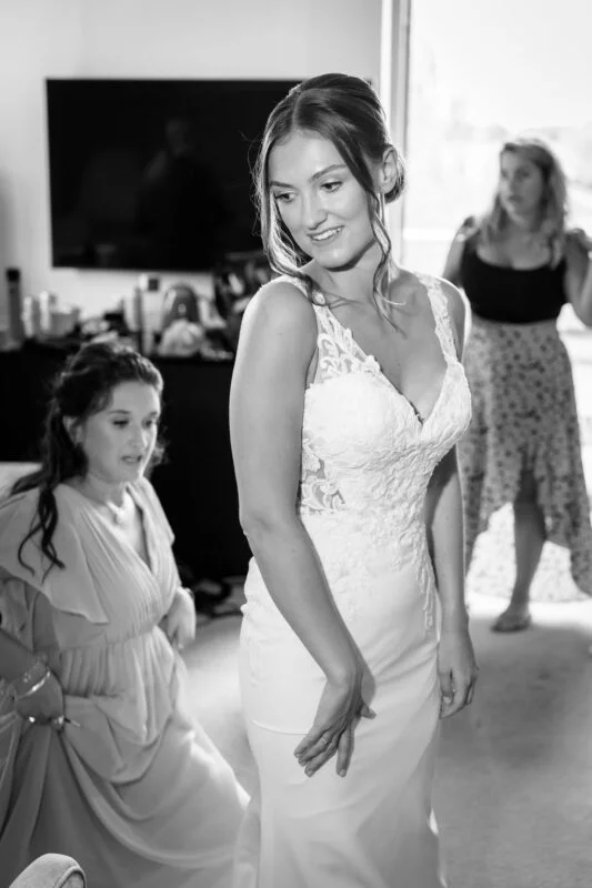 A glowing bride in a lace v-neck wedding dress smiles during bridal preparations while bridesmaids assist in the background.
