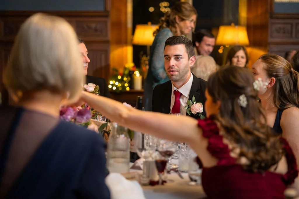 A groom in a black suit and red tie sits at a dinner table during a wedding reception, surrounded by guests in a warmly lit panelled room.