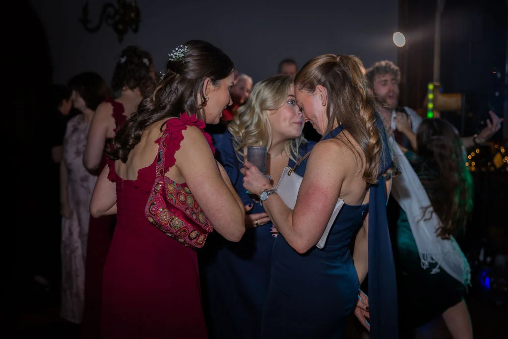 Wedding guests in elegant evening dresses converse closely together in a dimly lit reception room filled with celebratory energy.