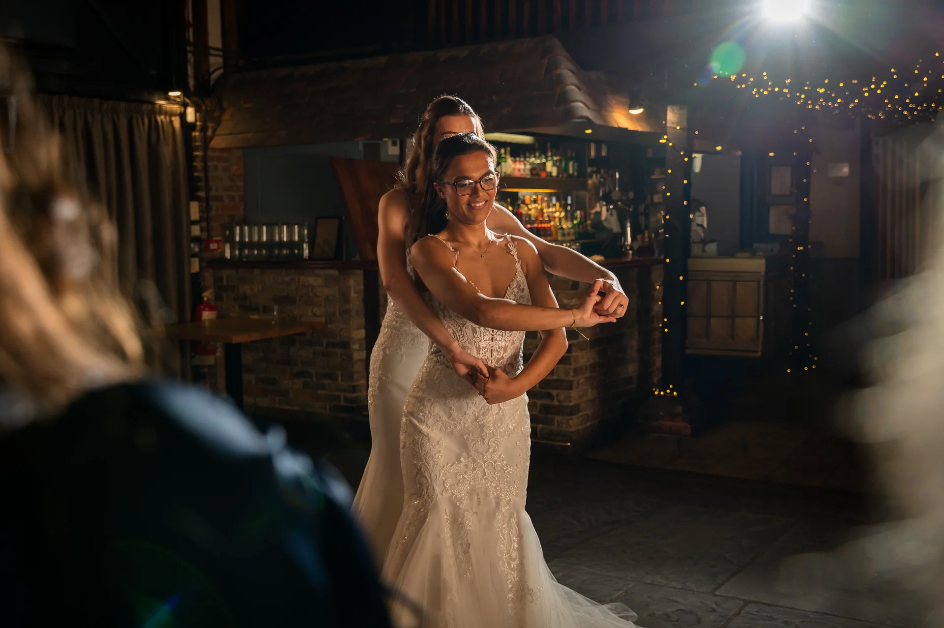 Two brides share a joyful first dance together at an evening wedding reception inside a cosy pub venue illuminated by string lights.