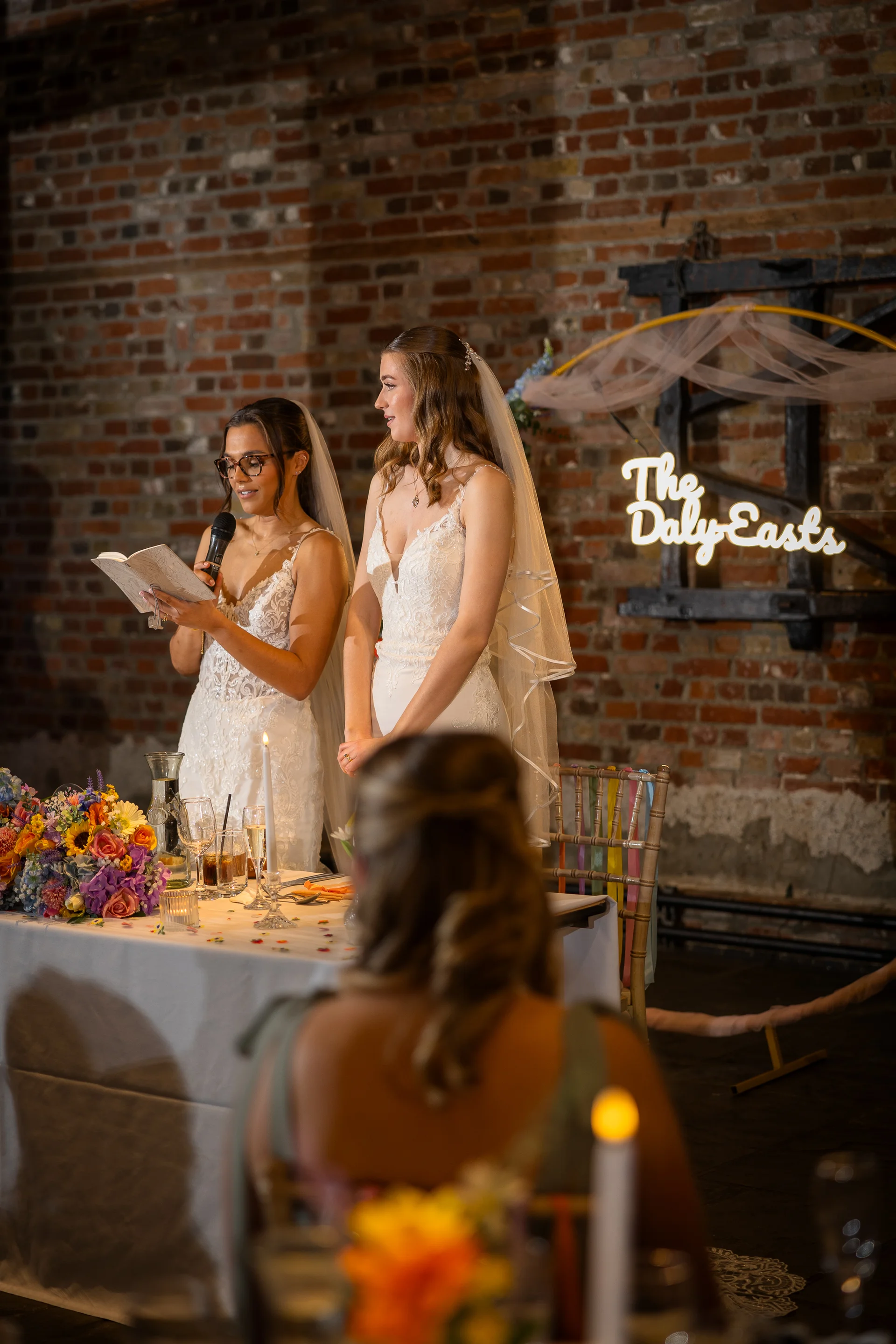 Two brides deliver a speech at a wedding reception set against an exposed brick wall, with one bride holding a microphone and notes while the other stands smiling beside her.