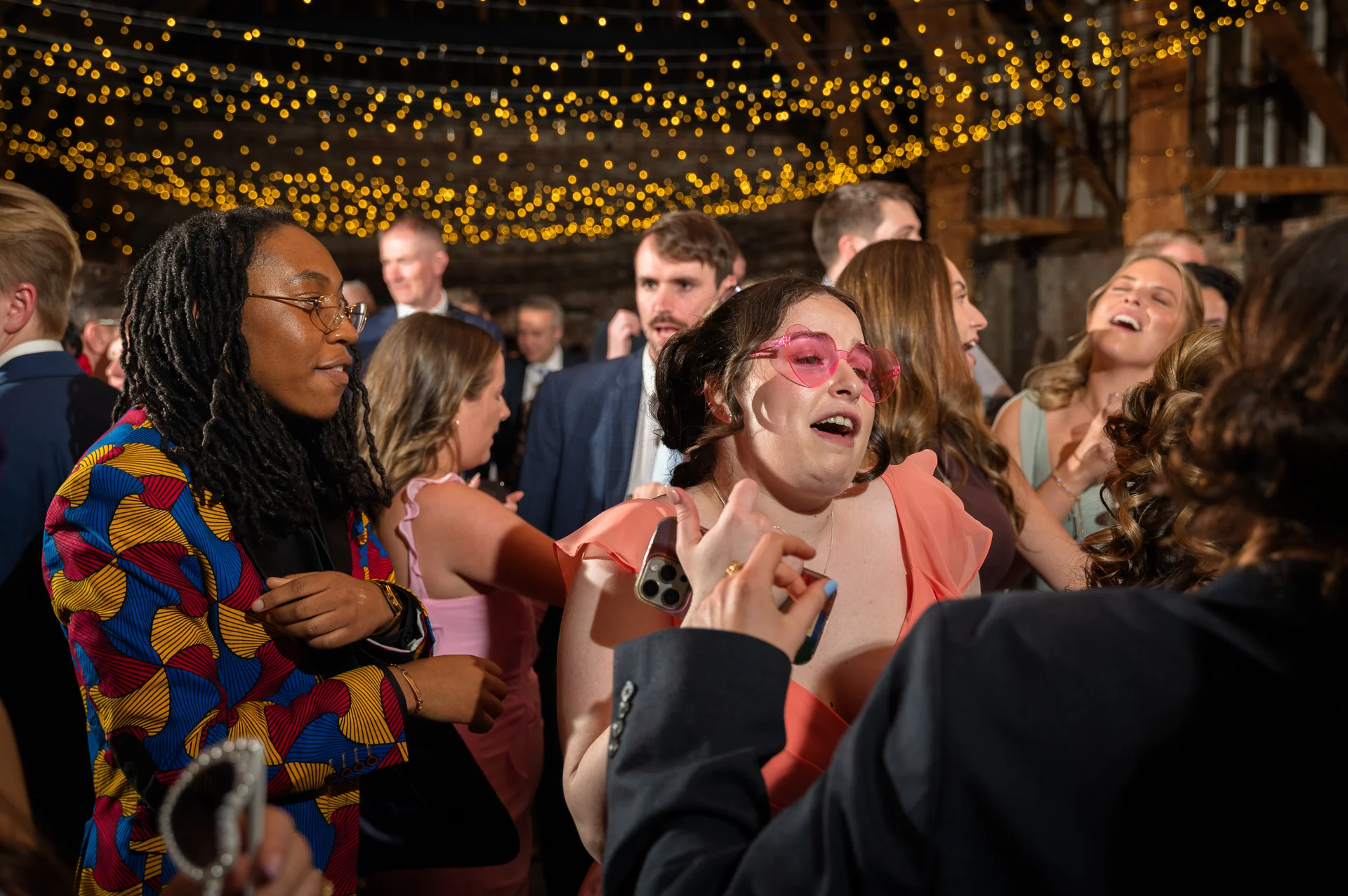 A joyful group of wedding guests dancing and laughing under a warm fairy light canopy inside a rustic barn venue.