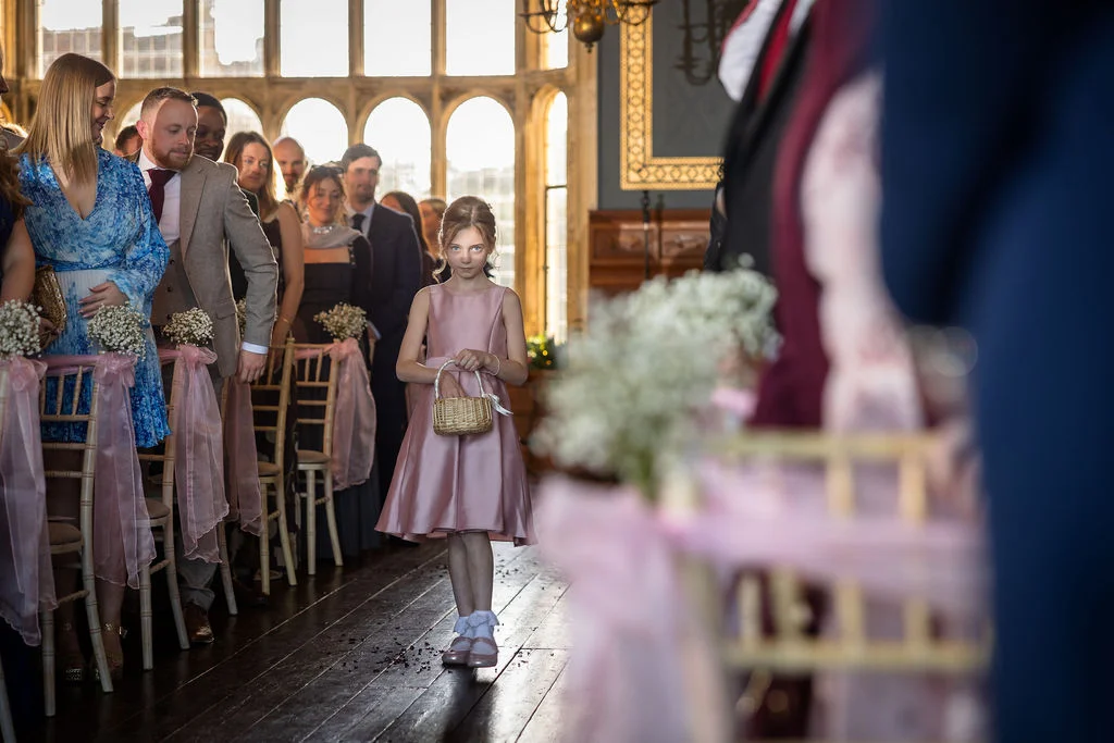 A young flower girl in a pink satin dress walks down the aisle holding a small wicker basket during an indoor wedding ceremony.