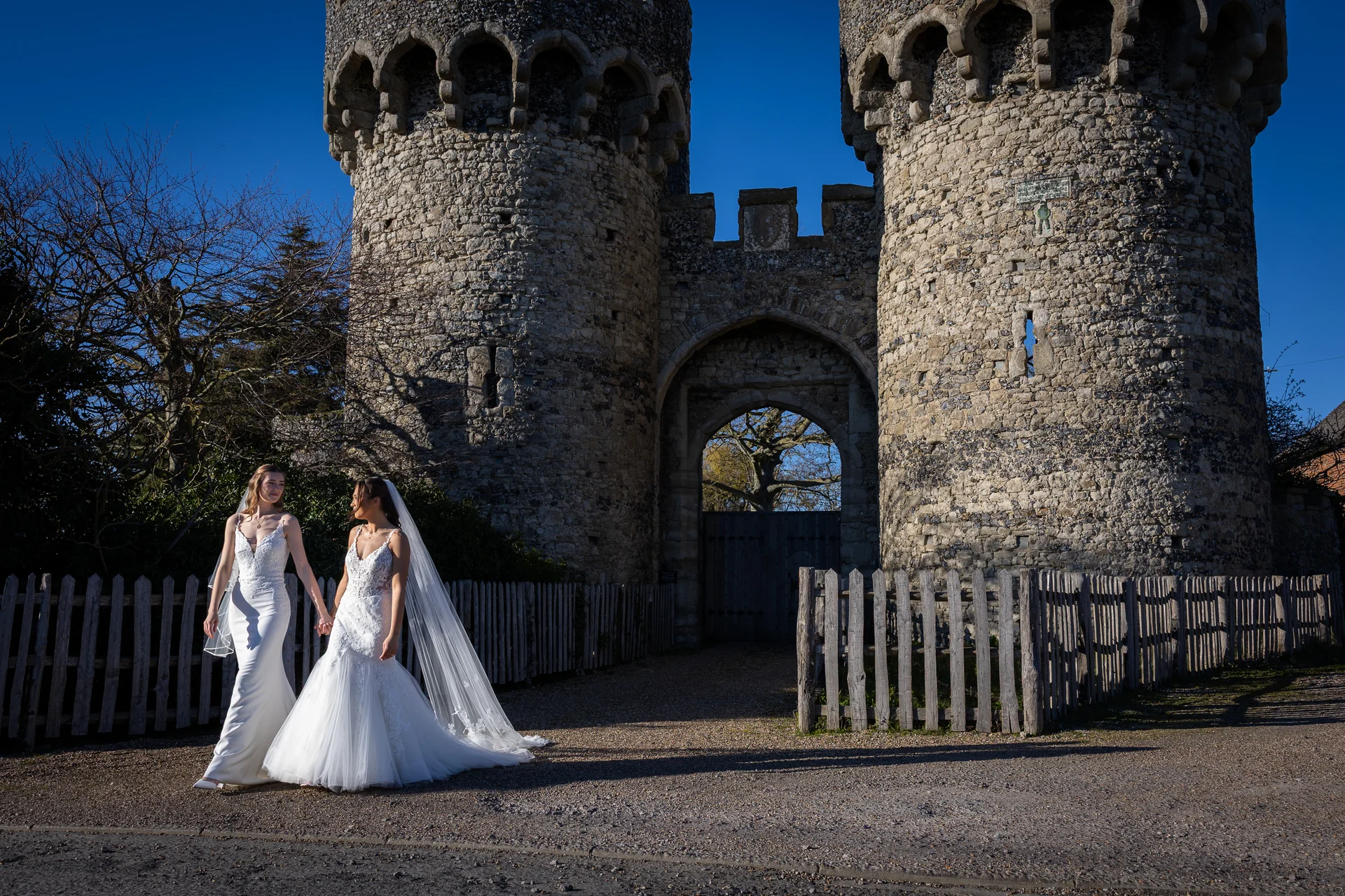 Two brides in white wedding dresses walk hand in hand in front of a historic stone castle gate under a bright blue sky.