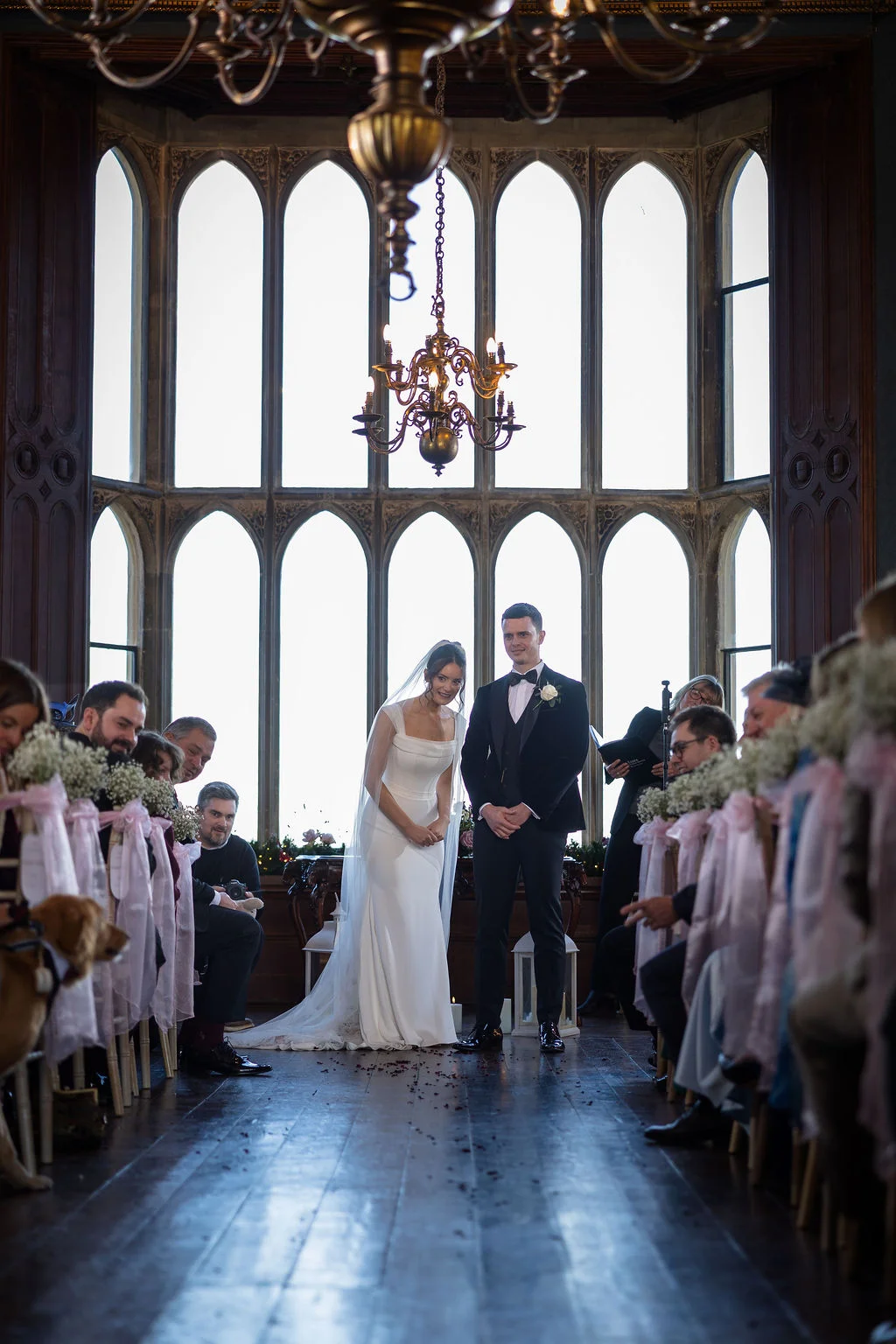 A newly married couple stands together at the end of the aisle inside a grand manor house with large arched windows and guests seated on either side.