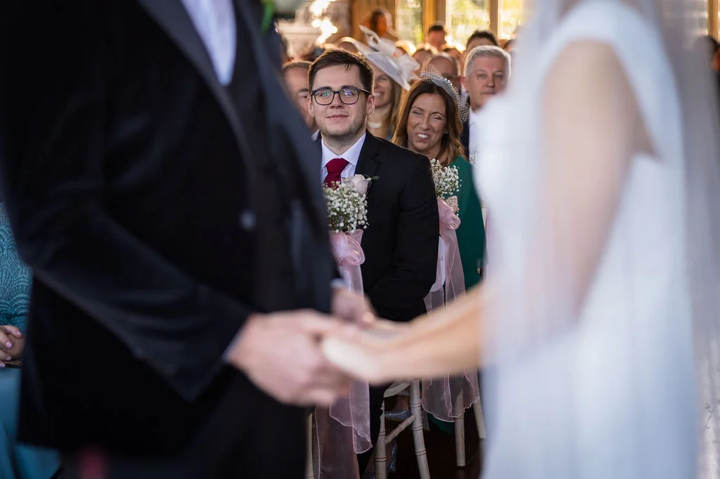 An over-the-shoulder view of a bride and groom holding hands during their wedding ceremony, with wedding guests watching and smiling in the background.