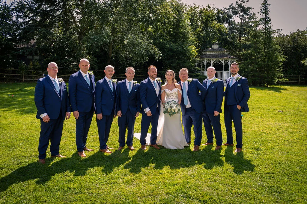 A newlywed bride standing in a white gown and holding a bouquet is surrounded by a large group of groomsmen in matching navy suits on a sunny lawn with a gazebo in the background.
