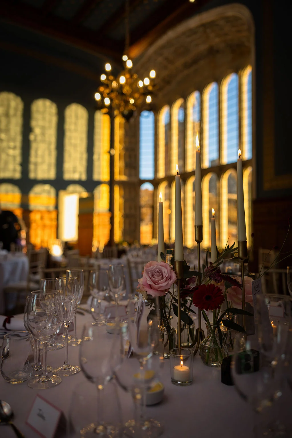 An elegantly set wedding reception table features taper candles in tall brass holders and bud vases with pink roses inside a grand hall with towering arched windows.