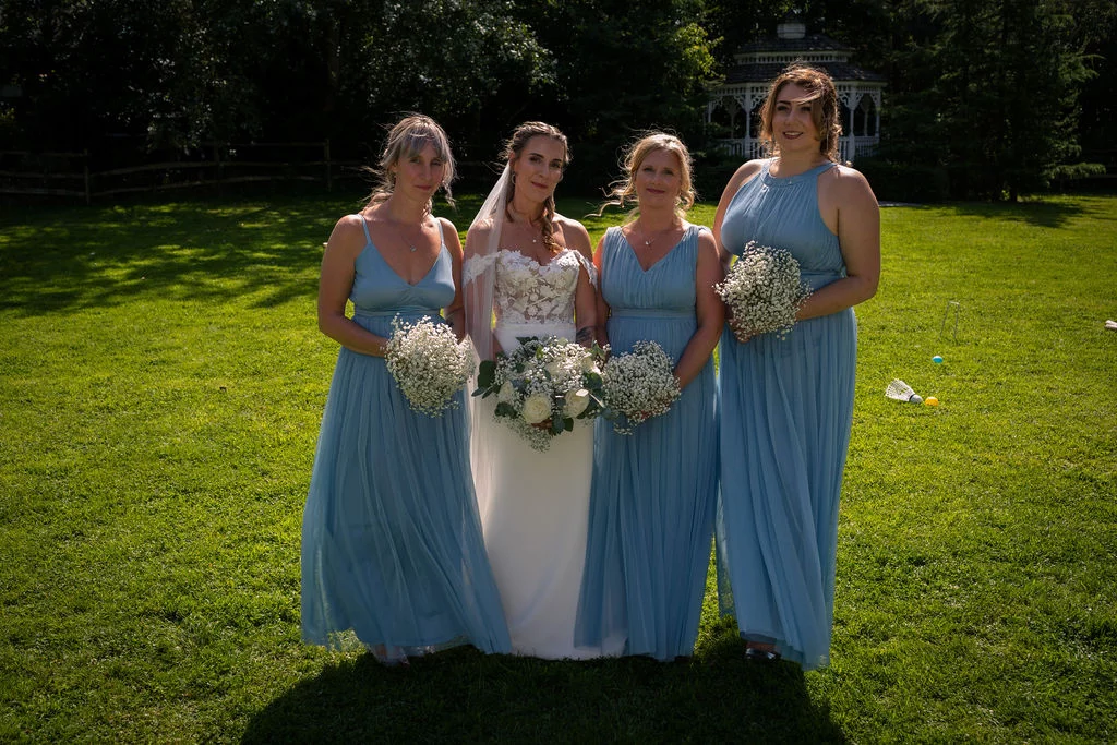 The bride stands in a white gown and veil holding a lush bouquet, flanked by four bridesmaids wearing matching dusty blue chiffon dresses on a sunny lawn.