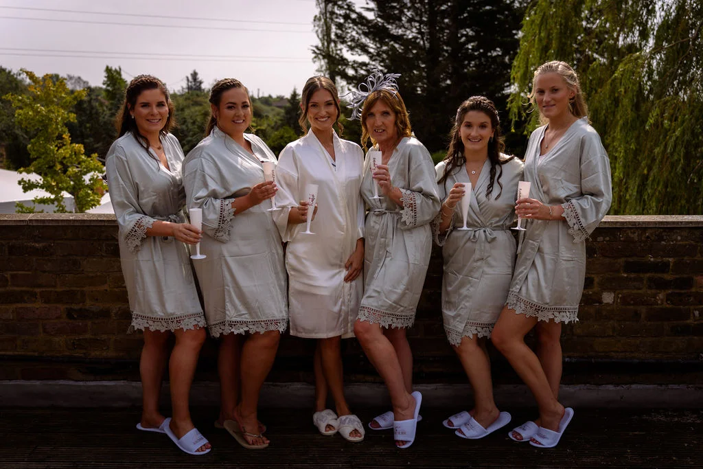 The bride in a white robe stands smiling with her bridesmaids and mother wearing matching sage robes and holding champagne flutes outdoors against a brick wall.