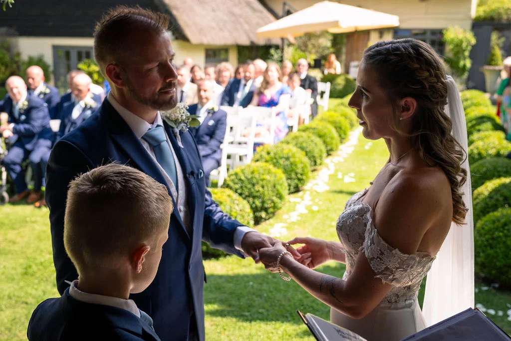 A groom and bride exchange rings during an outdoor garden ceremony while a young page boy looks on attentively.