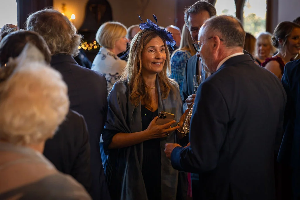 Wedding guests mingle and converse happily during a drinks reception, with a smiling woman wearing a blue fascinator as the focal point.