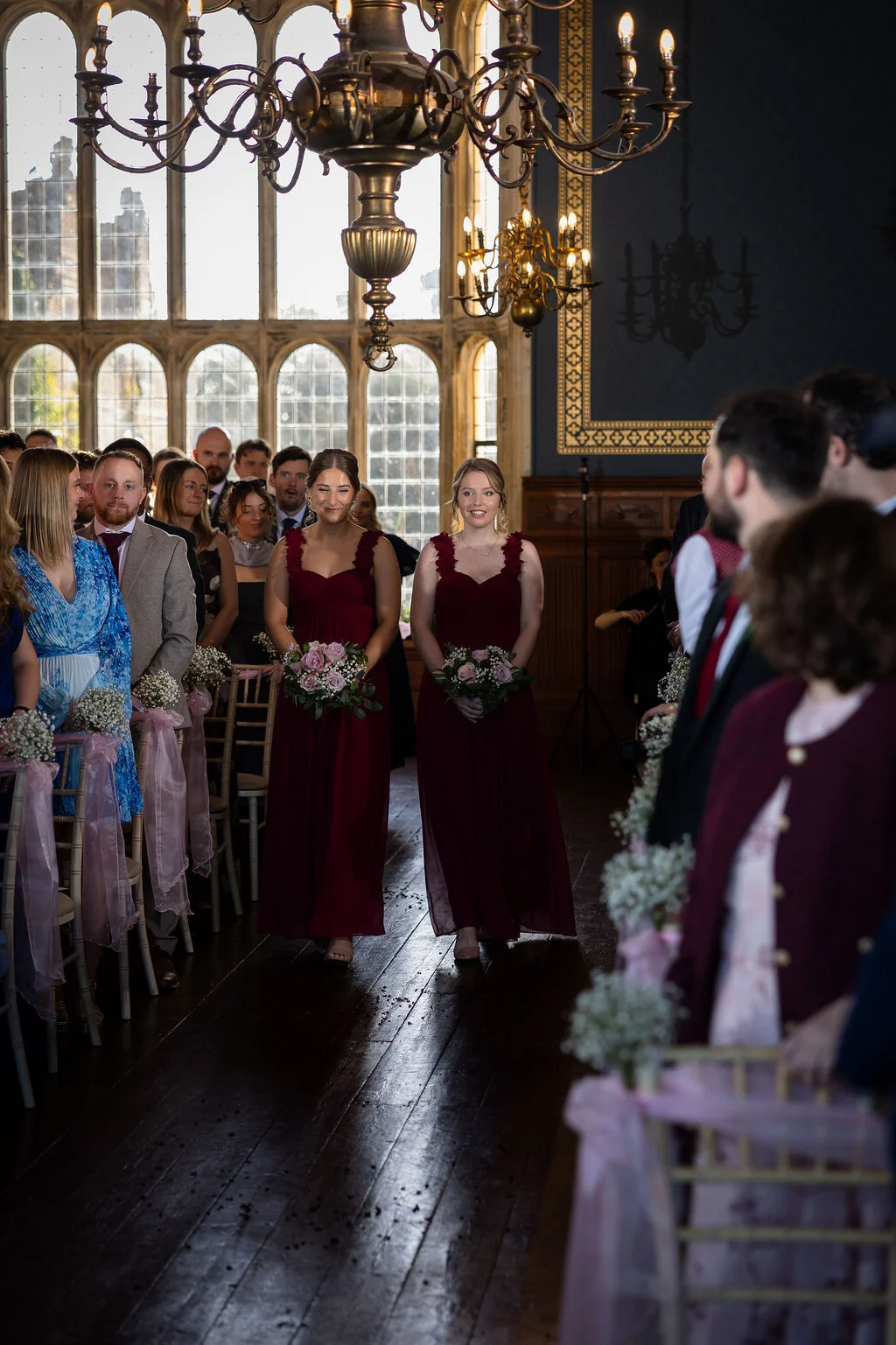 Two smiling bridesmaids in matching burgundy gowns holding bouquets walk down the wooden aisle of a stately home ceremony venue.