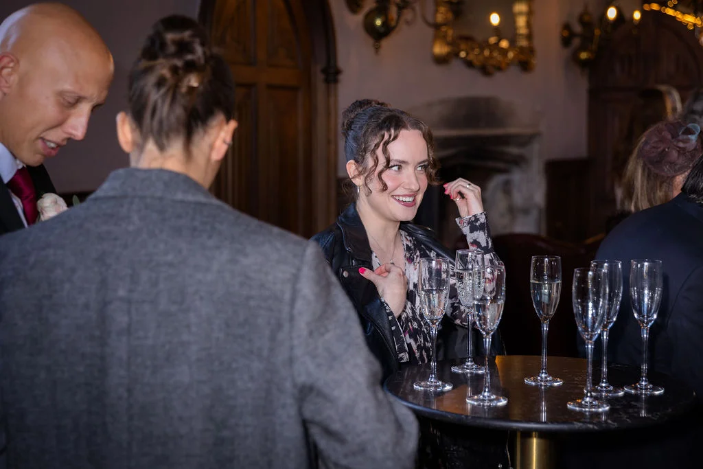 Wedding guests mingle and smile while standing around a high table set with champagne flutes during an indoor drinks reception.