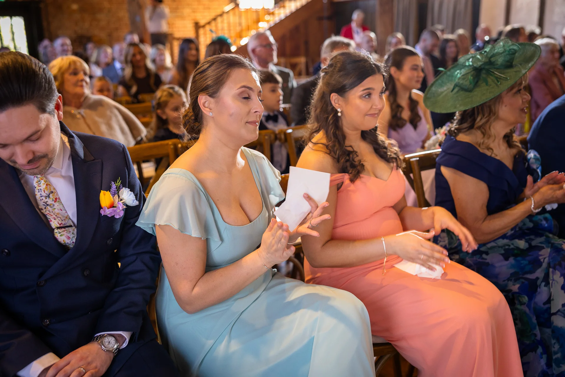 A candid photograph of wedding guests sitting in a rustic barn venue, with bridesmaids dressed in sage and pink gowns smiling and clapping during the ceremony.