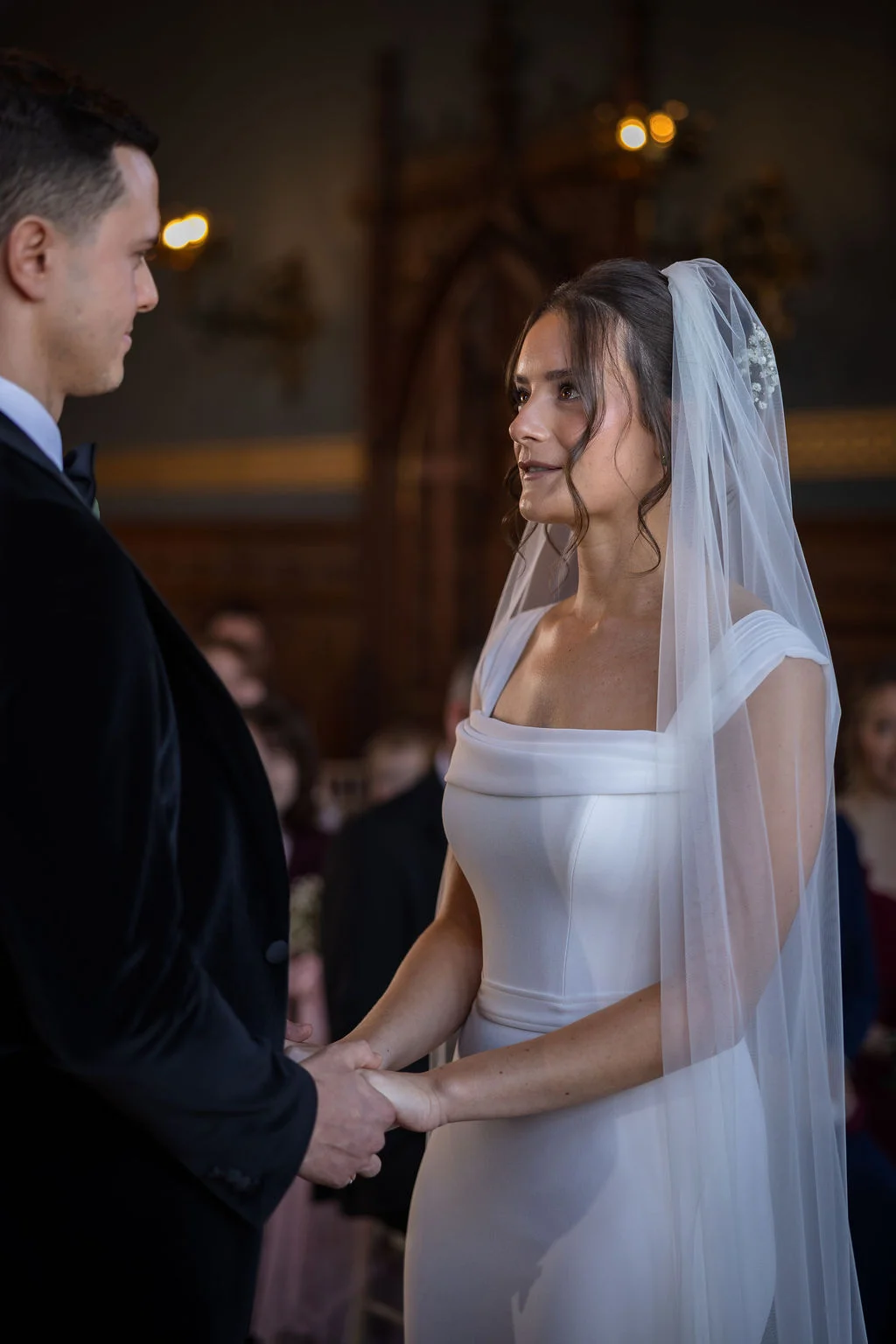 A bride and groom share a tender moment holding hands and looking into each other's eyes during their indoor wedding ceremony.