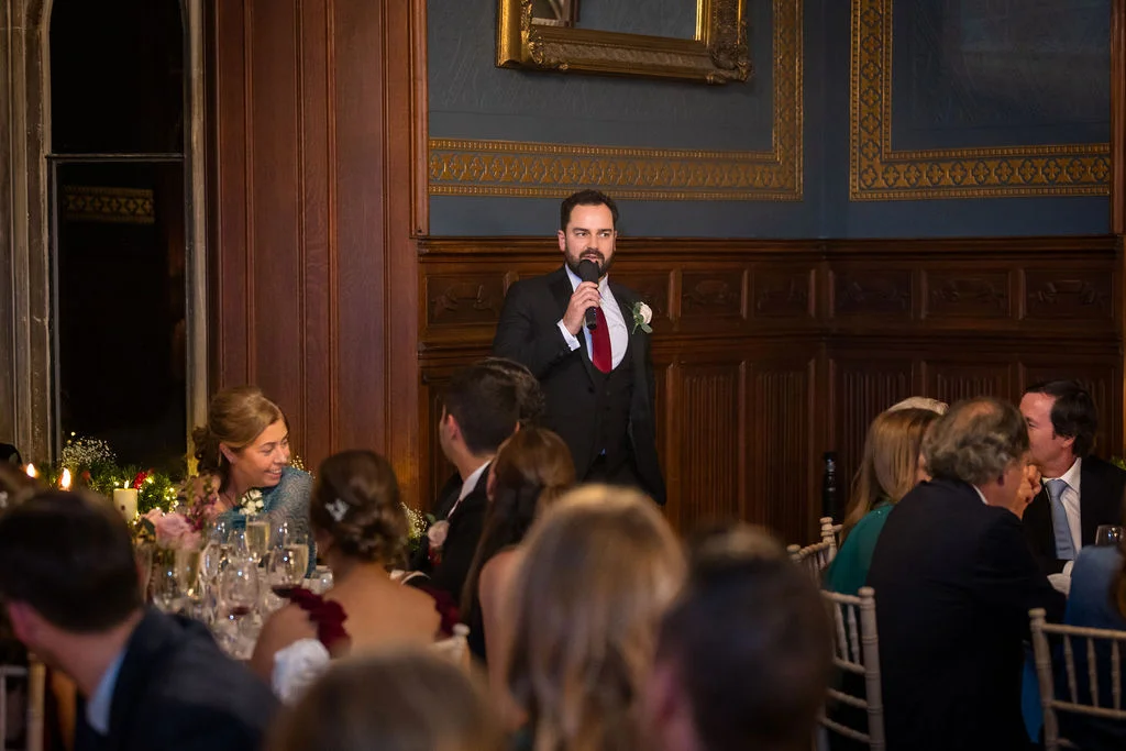 A groom delivers a heartfelt speech holding a microphone inside an elegant wood-panelled reception room filled with seated wedding guests.