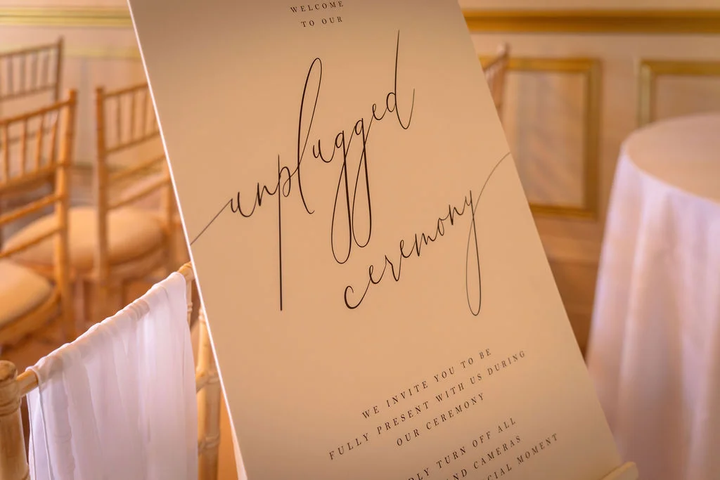 A modern unplugged ceremony sign stands on an easel inside a bright venue with gold chairs.