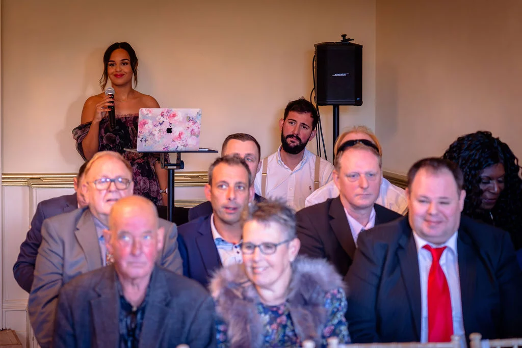 A smiling bride delivers a speech holding a microphone, standing behind a floral laptop at a wedding reception while attentive guests listen.
