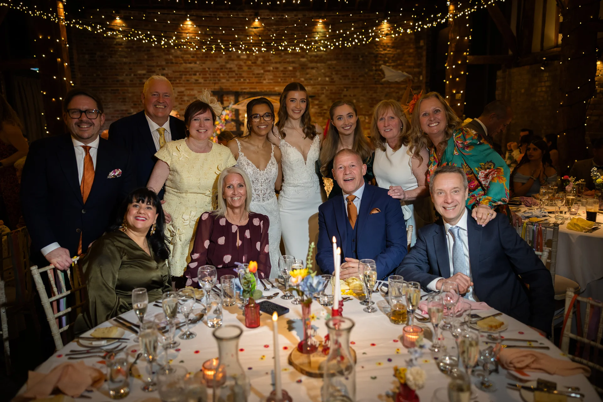 A large group of wedding guests and family members smile together around a banquet table inside a rustic barn decorated with fairy lights.