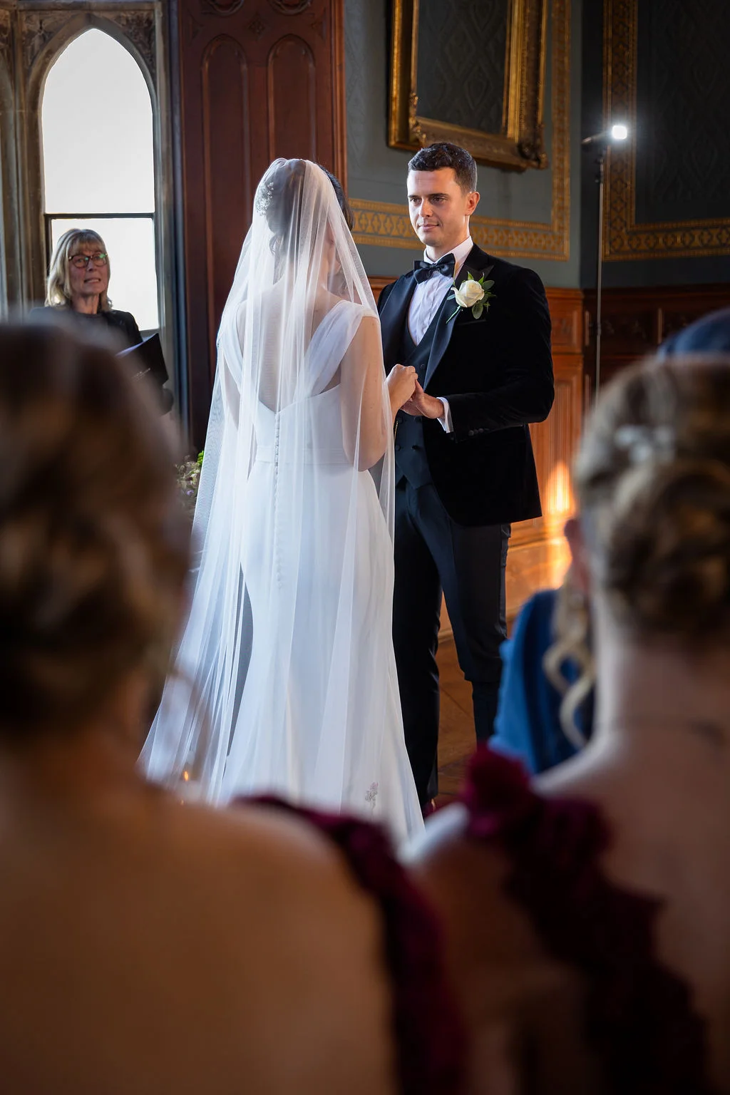 A bride and groom stand holding hands during their wedding ceremony inside a historic venue framed by guests in the foreground.