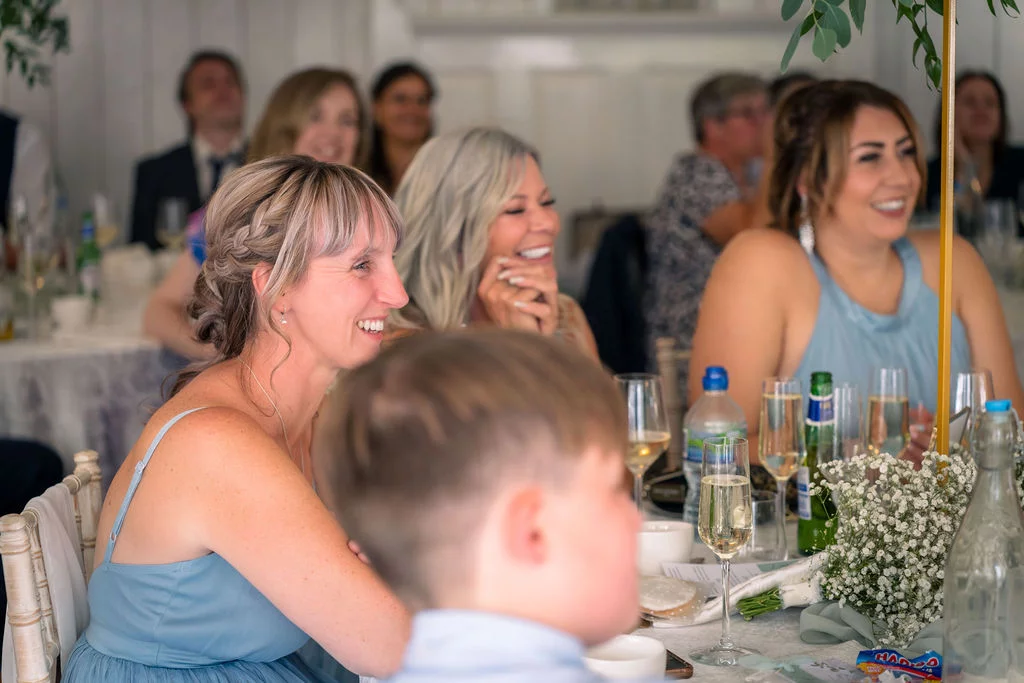 Wedding guests in blue attire smile and laugh together during the wedding breakfast seated at a table adorned with gypsophila florals.