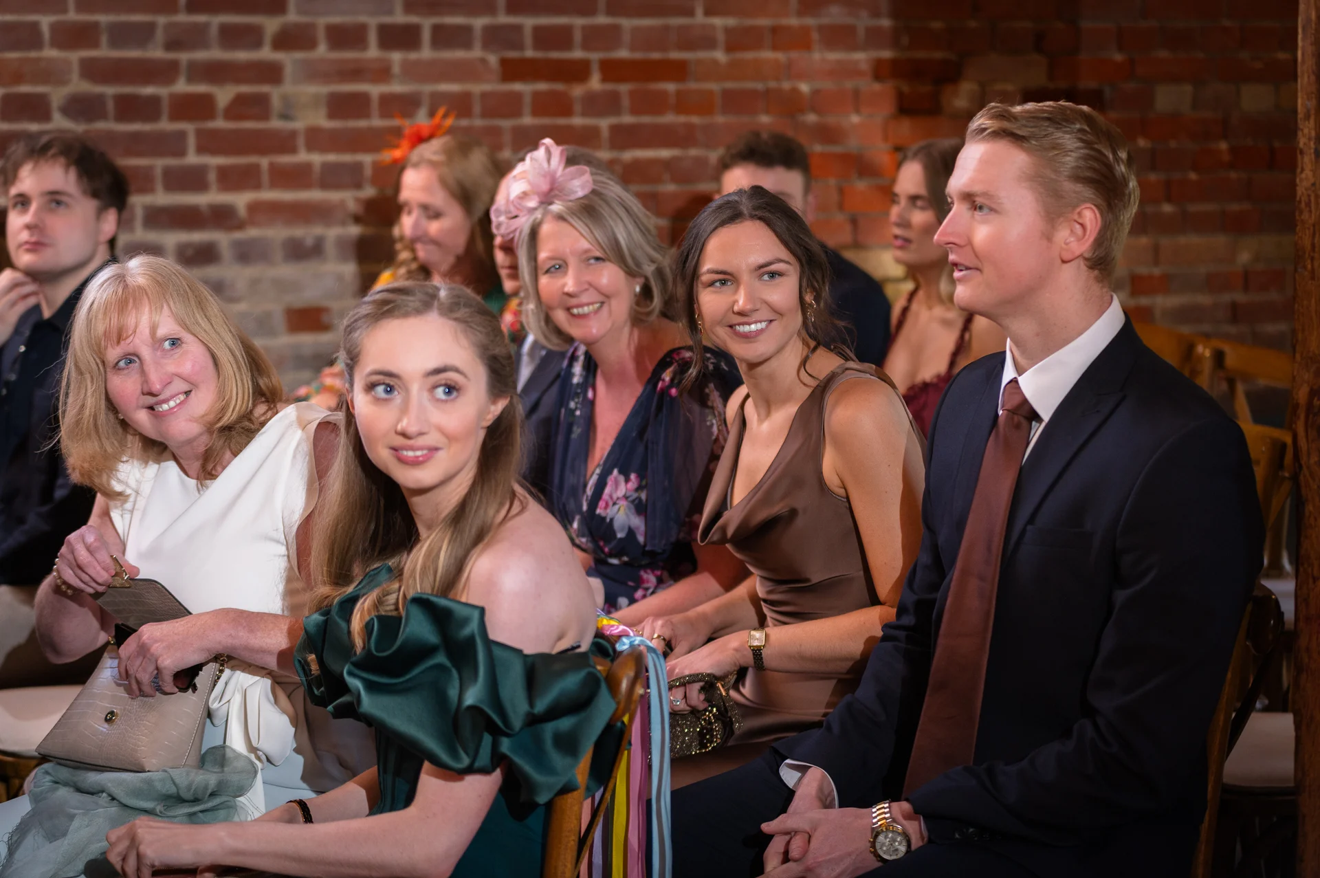 A candid shot of smiling wedding guests seated together in a rustic venue featuring an exposed brick wall background.