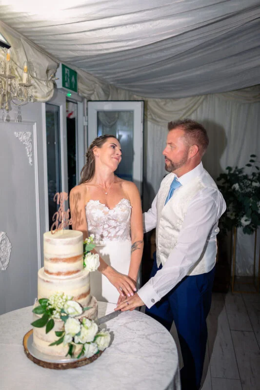 A smiling bride and groom cut into a three-tier semi-naked wedding cake adorned with white roses inside a marquee draped in white fabric.