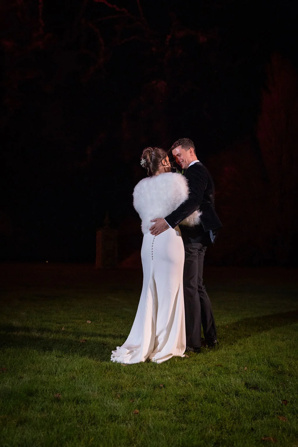 A newlywed couple embraces lovingly on a dark lawn at night, illuminated by soft lighting with the bride wearing a white crepe gown and a fluffy stole.