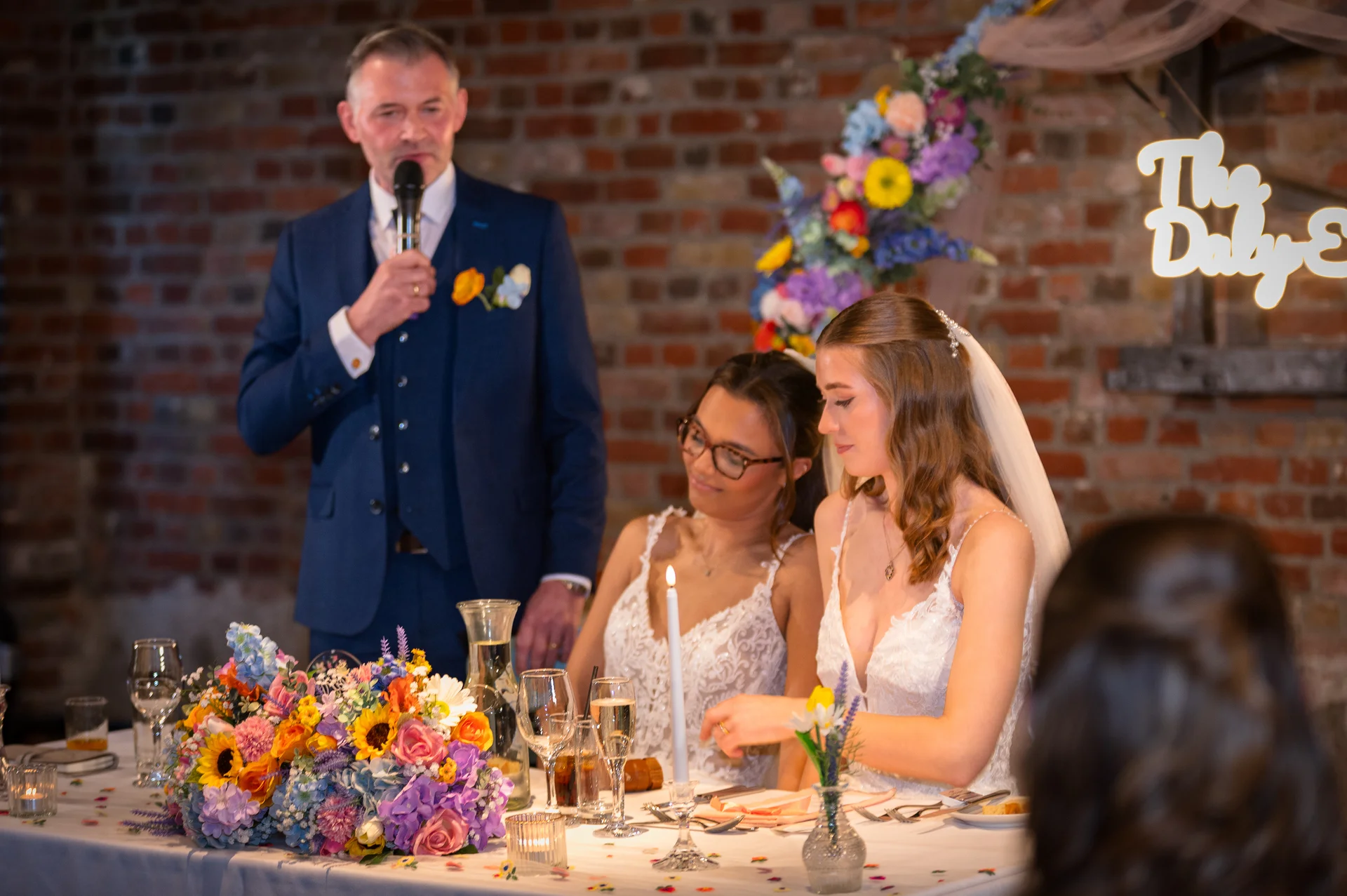 A guest gives a speech at the top table while two brides in lace wedding dresses sit together smiling in front of a rustic exposed brick wall.