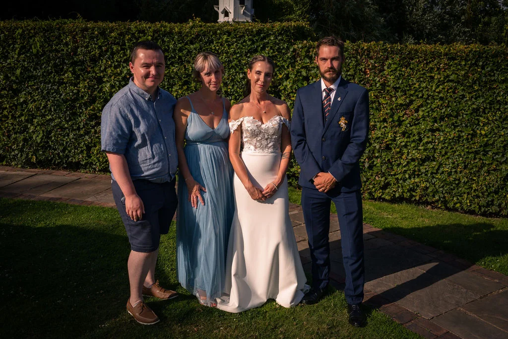 A bridal party and guests pose for a formal group portrait outside against a tall green hedge during the golden hour.