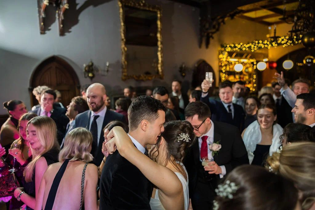 A newlywed couple share an intimate embrace on a crowded dance floor during their evening reception inside a historic castle venue adorned with warm fairy lights.