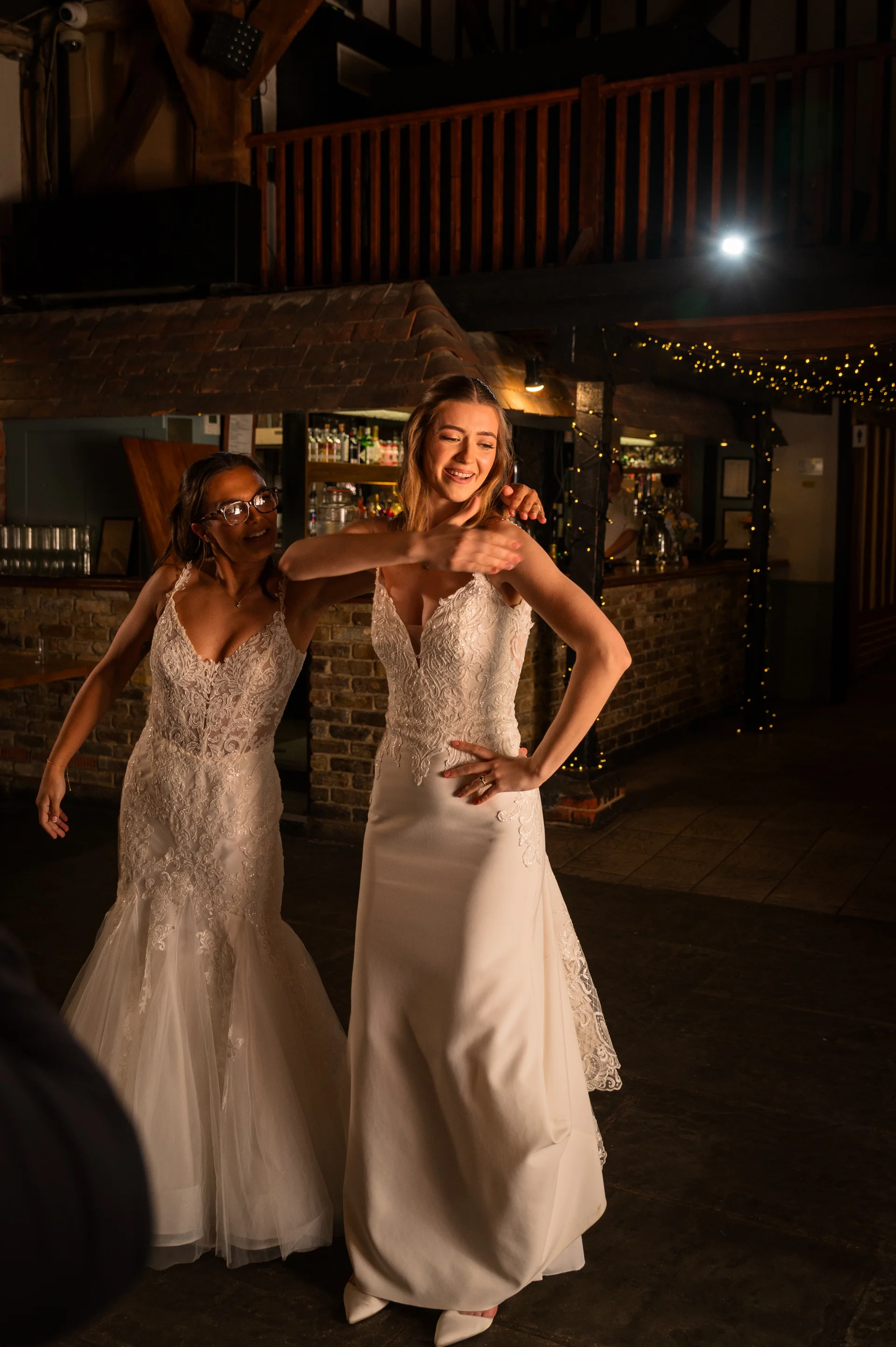 Two smiling brides in lace and satin wedding gowns dance happily together during an evening reception inside a rustic stone barn.