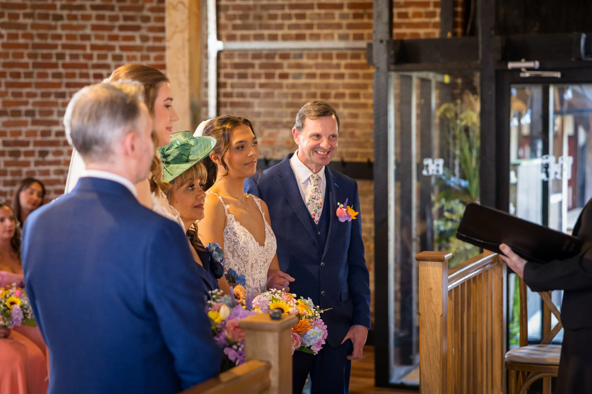 The bride and groom stand together during a civil ceremony inside a rustic brick barn venue, flanked by family members and guests.