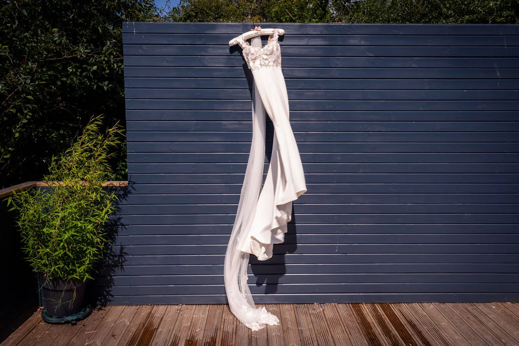 A elegant wedding dress with lace bodice and long veil hangs against a modern dark blue slatted wooden wall outside in bright sunlight.