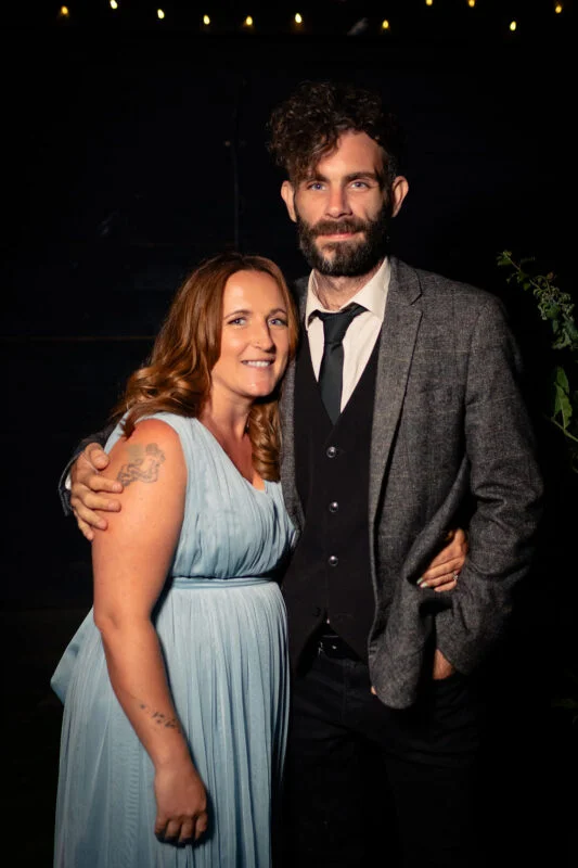 A smiling couple poses together outdoors at night, with the groom wearing a tweed jacket and waistcoat and the bride in a pleated dusty blue dress.