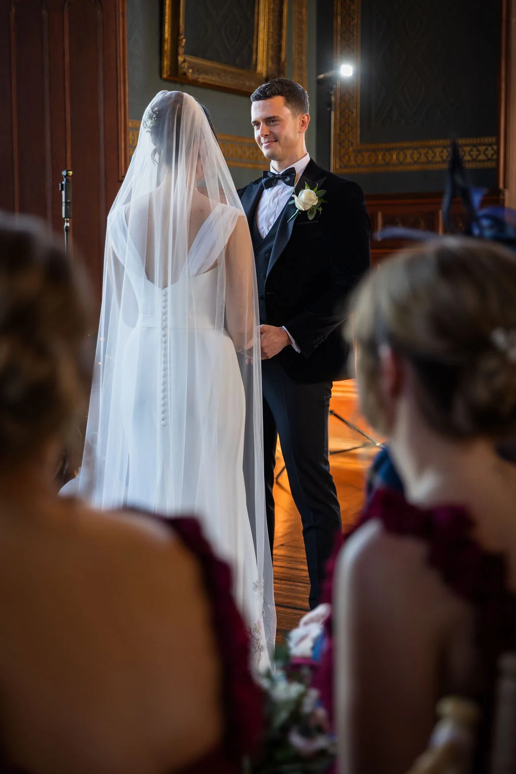 A bride in a sleek gown with a button-backed veil and a groom in a black velvet tuxedo stand together at the altar, framed by guests in the foreground.