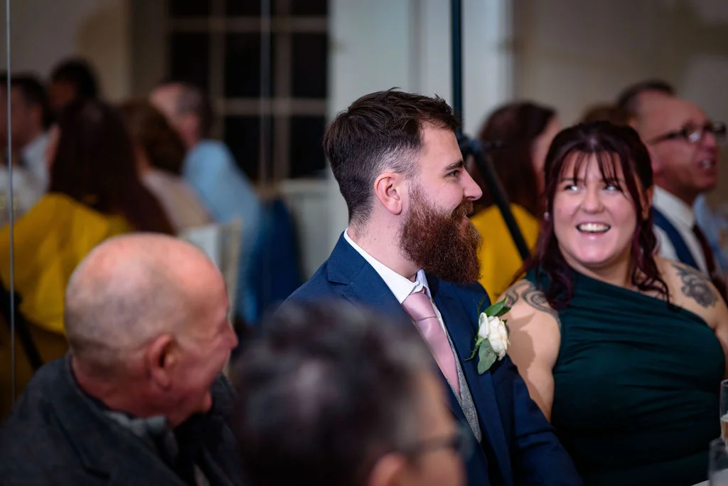 A groom with a beard and a navy suit laughing during the wedding speeches alongside smiling guests.
