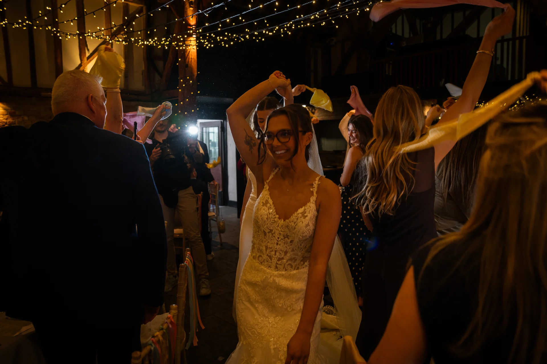 A smiling bride wearing glasses and a lace gown dances indoors beneath a twinkling fairy light canopy during an evening barn wedding celebration.