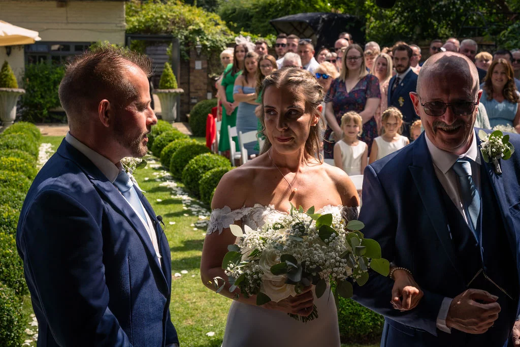 A glowing bride walks down a sunlit outdoor garden aisle on her father's arm, approaching the waiting groom who smiles warmly.