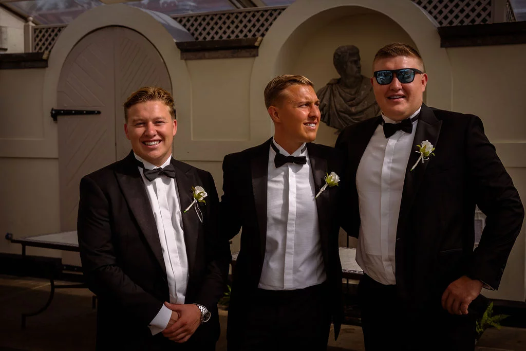 Three groomsmen in matching black tuxedos and bow ties stand together outdoors, smiling and laughing in the bright sunshine.