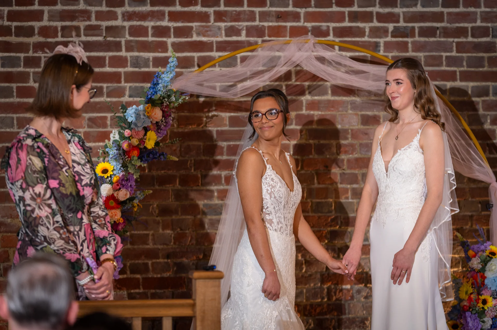 Two brides hold hands during an indoor ceremony in front of an exposed brick wall decorated with a floral arch and draped fabric.